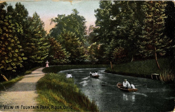 Historical postcard of Fountain Park in Piqua, Ohio. On the left side, is a walking path lined with trees. The right side shows a waterway where people are rowing boats. There are trees along the waterway and in the background.